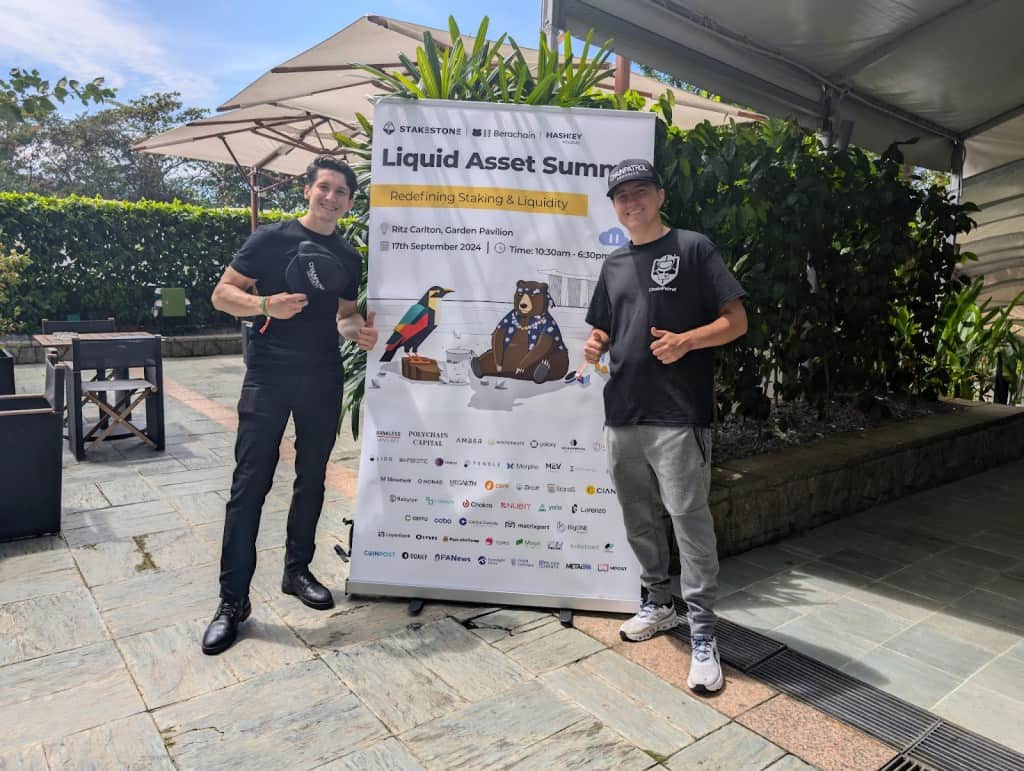 two men wearing black shirts standing in front of a sign that reads "liquid asset summit" while giving a thumbs up